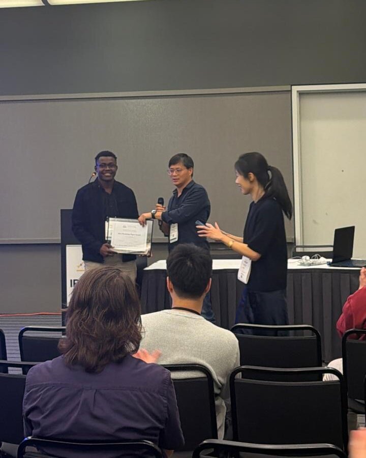 Austine holding the Best Paper Award certificate on stage with the workshop organizers