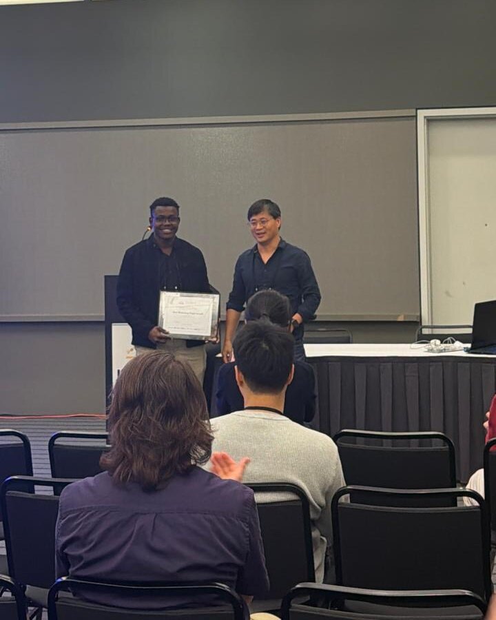Austine smiling and holding the award certificate beside the presenting organizer
