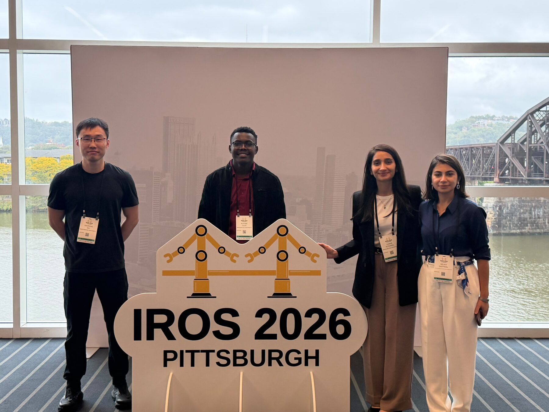Four Binghamton University researchers standing behind the IROS 2026 Pittsburgh sign with the city skyline behind them